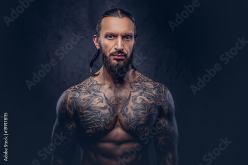 Close-up portrait of a muscular bearded tattoed male with a stylish haircut, isolated on a dark background.