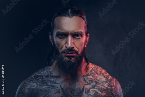 Close-up portrait of a muscular bearded tattoed male with a stylish haircut, isolated on a dark background.