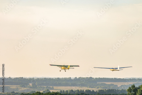 Small light-engine sport plane flying in the evening sky pulls on a rope the glider plane. Flight over the green hazy plain.