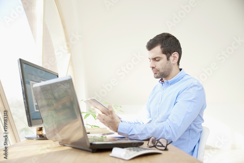 Professional man with digital tablet, laptop and personal computer. Portrait of young businessman looking thoughfully while sitting at office desk and using digital tablet. 