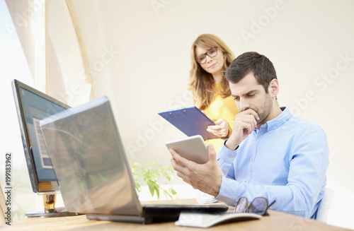 Business team working at the office. Young financial businessman sitting in front of laptop and using digital tablet while businesswoman consulting with him. 