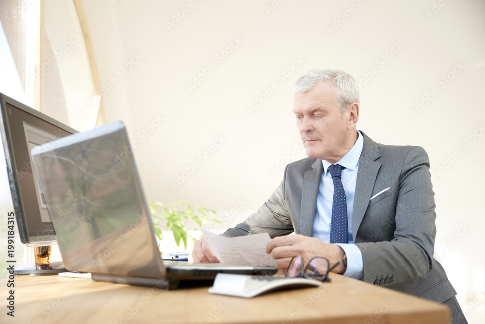 Portrait of an elderly businessman wearing suit while sitting at office desk in front of laptop. Portrait of an elderly businessman wearing suit while sitting at office desk in front of laptop.
