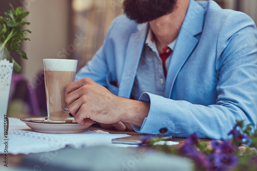 Portrait of a fashionable bearded male with a stylish haircut, holds a glass of a cappuccino, sitting in a cafe outdoors.