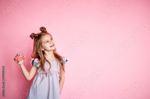 fashionable little girl with long brunette hair in pink dress holding a perfume, isolated on a pink background with copy space