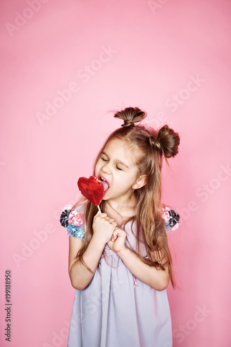 funny little girl with long brunette hair in dress, licks tongue lid in the form of heart in hands, isolated on pink background. 