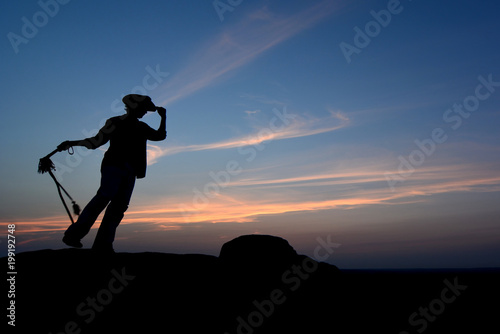 Stylish cowboy at sunset standing on rock and cracking whip. Male silhouette on romantic blue sky in mexico. Mans fashion concept like western film. 
