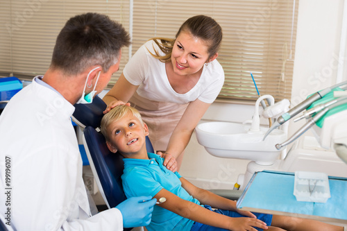 Young boy is sitting in dental chair and mother is supporting him