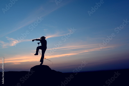 Cowboy crack whip in a jump on rock. Mexican scene of male silhouette on romantic cloudy blue sky background. 