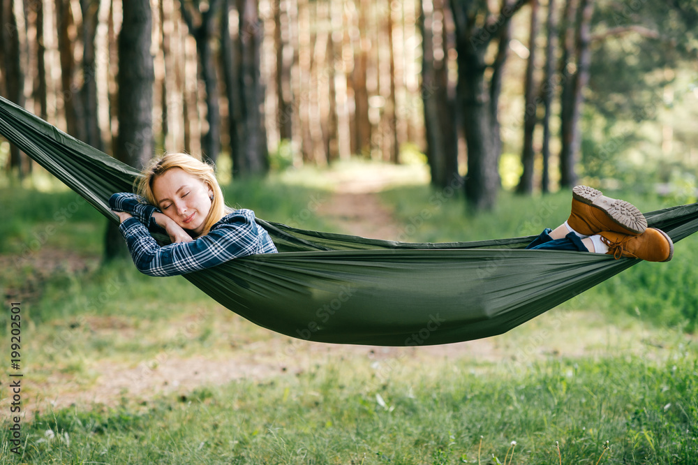 Outdoor lifestyle portrait of young beautiful blonde girl sleeping in  hammock in forest. Cute woman dreaming at nature in summer. Tired female  resting in camping trip after hard walk. Travel equipment Stock