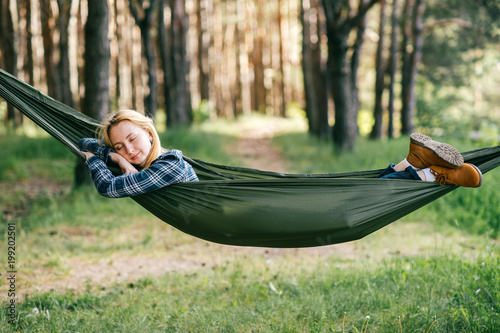 Outdoor lifestyle portrait of young beautiful blonde girl sleeping in hammock in forest. Cute woman dreaming at nature in summer. Tired female resting in camping trip after hard walk. Travel equipment