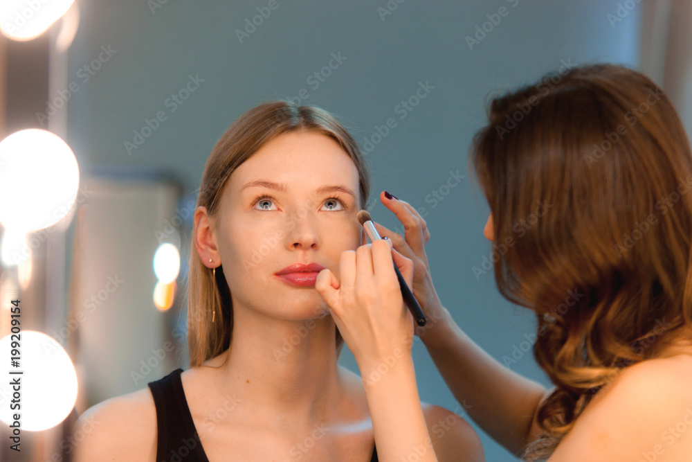 Makeup artist applies makeup to attractive blonde girl at the mirror with  lamps in the beauty studio. A satisfied customer of make-up and hairstyle  service. Work in beauty salon Stock Photo