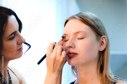 Makeup artist applies makeup with brush in hand to attractive blonde girl in the beauty studio.