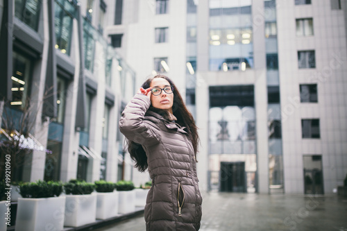 Portrait beautiful young business woman brunette in jacket and sweater stands on background office building, business center in glasses for view. Hand holding onto shackle of glasses and looking away