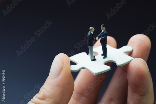 Business success strategy with collaboration, teamwork or negotiation jigsaw key, miniature people businessmen handshaking on white jigsaw puzzle piece in real human hand, dark black background photo