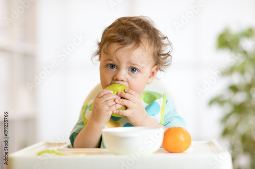 Cute baby boy one years old sitting on high children chair and eating fruits alone in white kitchen