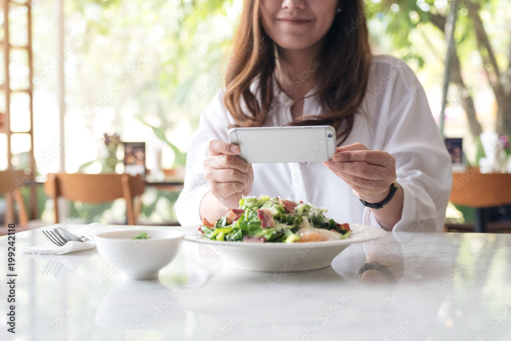 Closeup image of an asian woman using smartphone to taking photo of salad in a white plate on table in cafe Closeup image of an asian woman using smartphone to taking photo of salad in a white plate on table in cafe