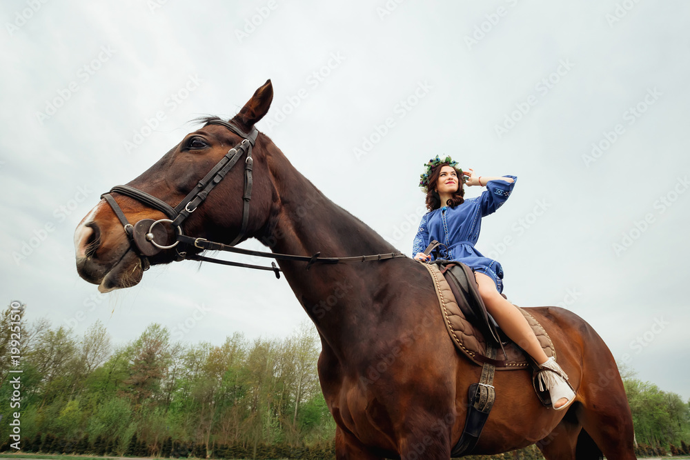 A beautiful girl with a wreath of flowers on her head sits in a saddle on a horse and looks distant A beautiful girl with a wreath of flowers on her head sits in a saddle on a horse and looks distant