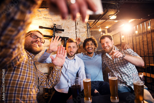 Canvas Print Four smiling multicultural friends waving and taking a selfie while sitting in the pub