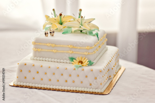 Two floor wedding white marchpane cake with flowers and wedding rings on top on gold metal plate square shape