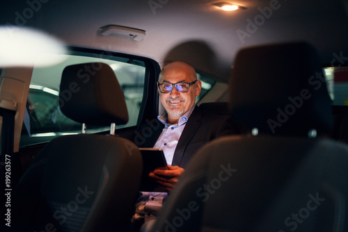 Mature professional happy successful businessman is being driven in the back seat of the car while using a tablet. © dusanpetkovic1