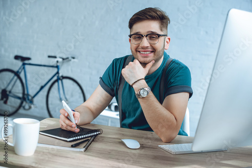 cheerful young man in eyeglasses smiling at camera while working at home office