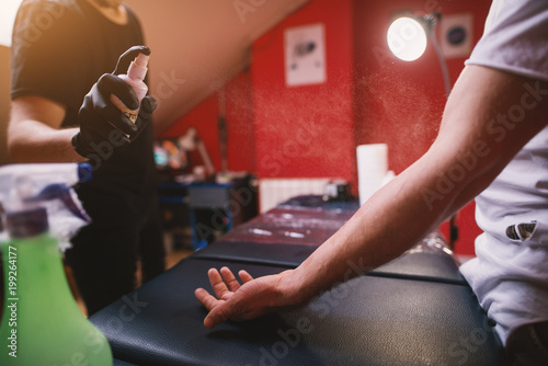 Tattoo artists spraying customers arm with spray for sterilization before further preparation for tattooing.