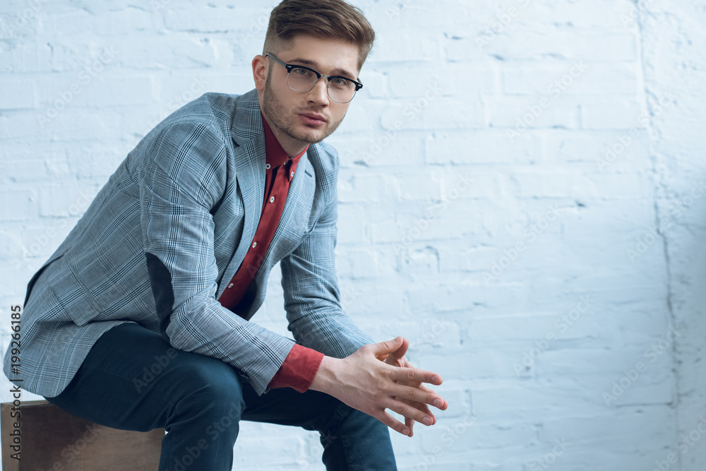 Attractive bearded man wearing suit and glasses and sitting in front of brick wall Attractive bearded man wearing suit and glasses and sitting in front of brick wall