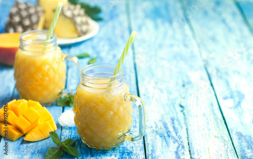Fresh pineapple and mango smoothie in two glasses with fruits on a torquise wooden rustic background