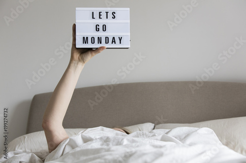Schilderij op canvas Female in bed under the sheets holding up a lets go monday sign