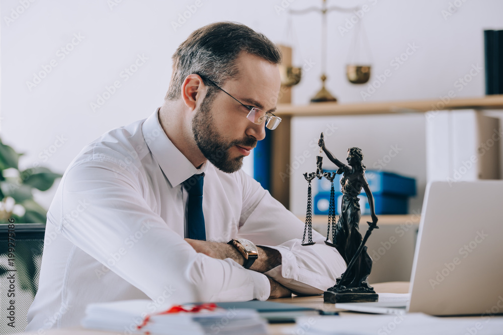 focused lawyer in eyeglasses looking at femida at workplace in office focused lawyer in eyeglasses looking at femida at workplace in office