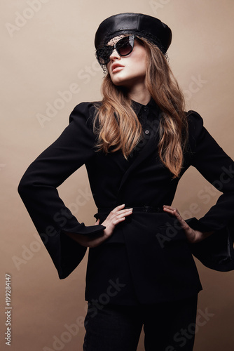 Fashion portrait of beautiful young woman in black flared jacket, black leather beret cap and dark sunglasses with veil. Standing with hands on waist
