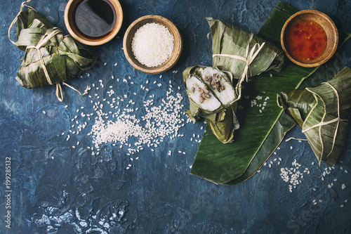 Asian rice piramidal steamed dumplings from rice tapioca flour with meat filling in banana leaves. Ingredients and sauces above over blue texture background. Top view, space. photo