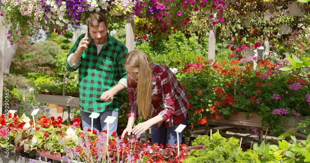 Relaxed casual man speaking with client on smartphone working with young woman in blooming garden selling flowers together. Relaxed casual man speaking with client on smartphone working with young woman in blooming garden selling flowers together.