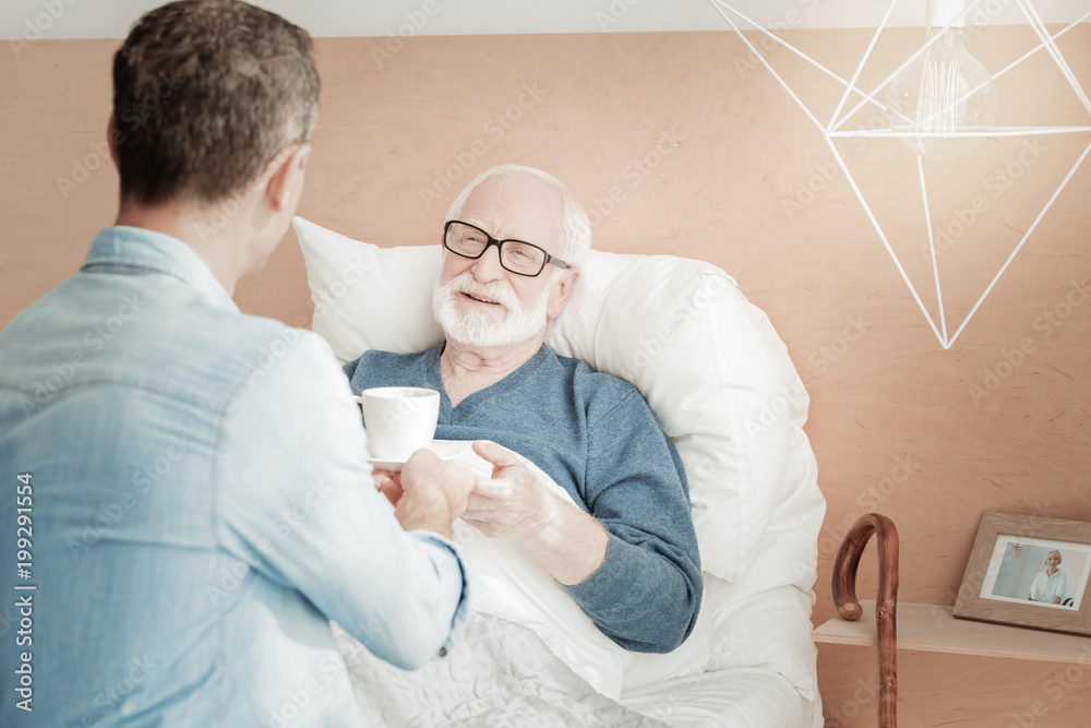 Fototapeta premium Reliable support. Satisfied grateful unshaken man being ill in the bedroom smiling and taking a cup.