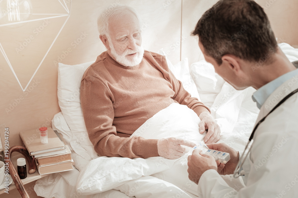 Fototapeta premium New pill. Tired senior unshaken man sitting on the bed listening to a doctor and looking at pills.