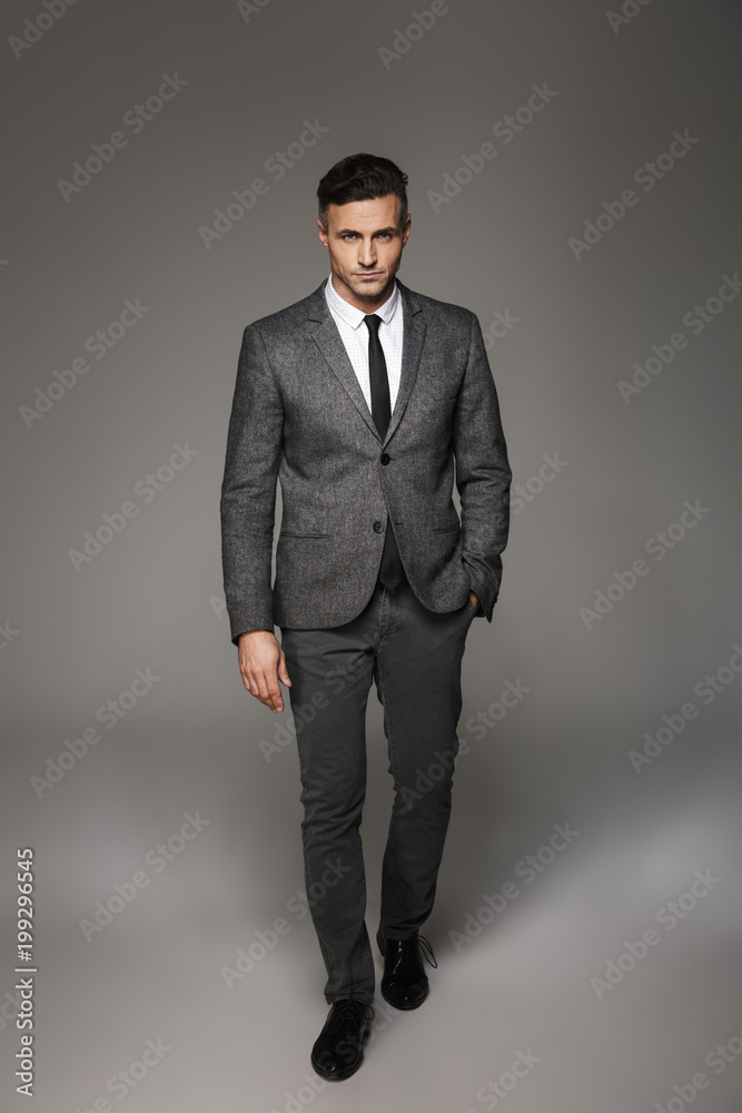 Full length portrait of successful businessman wearing business suit posing on camera with serious look and hand in pocket, isolated over gray background Full length portrait of successful businessman wearing business suit posing on camera with serious look and hand in pocket, isolated over gray background