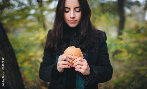Cute girl in a coat eating a hamburger while walking in an autumn forest. Fast food and hunger concept.