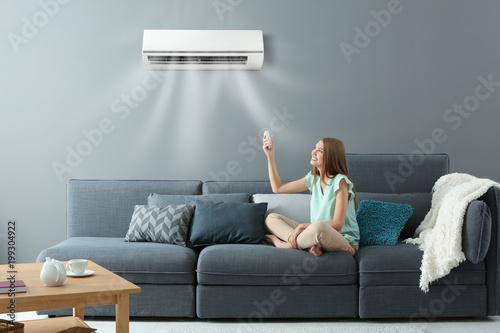 Young woman switching on air conditioner while sitting on sofa at home photo