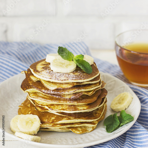 fresh baked pancakes with banana in white plate and cup of tea on blue linen on white background