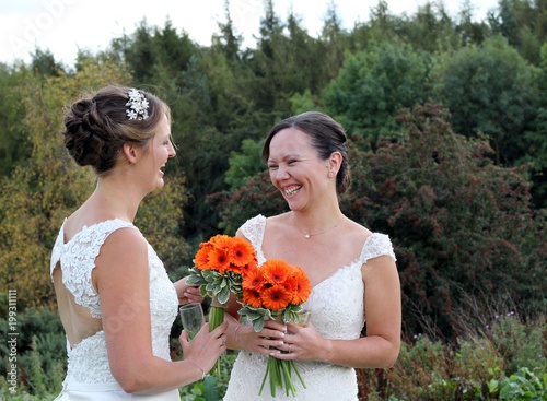 Brides on their wedding day, two smiling women looking happy and in love, holding orange gerbera bouquets , outdoors for a rural celebration, same sex occasion 