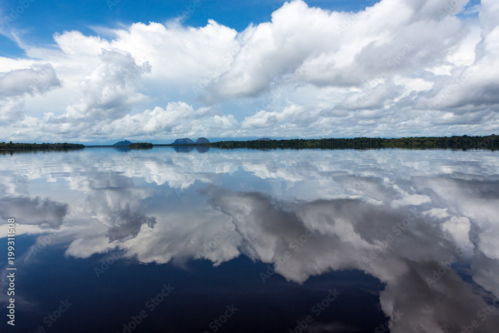 Obraz premium Beautiful reflection of blue sky and clouds in the waters of the Negro River with rainforest and hills in the background.