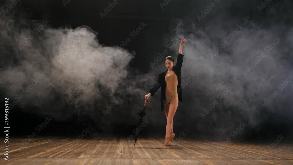 Ballerina in black long jacket on sexy bodysuit dancing on stage with magic light and smoke, slow motion. Young attractive dancer in ballet shoes pointe with umbrella. Ballerina in black long jacket on sexy bodysuit dancing on stage with magic light and smoke, slow motion. Young attractive dancer in ballet shoes pointe with umbrella.