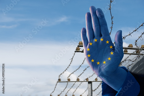 Hand with European flag stops immigration of refugees, blurred border fence in the background, blue sky with copy space