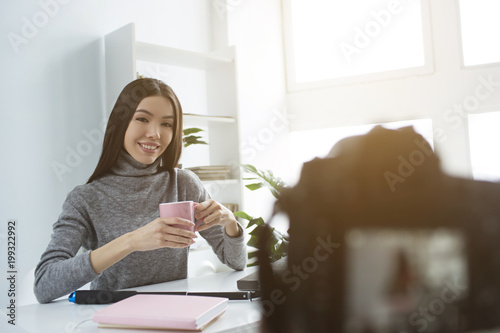 Nice and attractive girl is sittingat the table and drinking tea from pink cup. She is doing that in front of camera that recording it. She is looking to it and smiling.