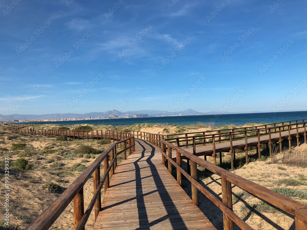 perspective view of some wooden walkways that form a path to the beach. In the distance two people come walking down the catwalks. In the distance, two perch rods are also almost imperceptible.