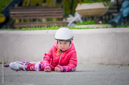 A sad little girl lies on the asphalt in a helmet and protective equipment. The child fell on roller skates. Unsuccessful roller skating