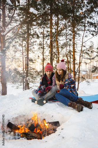 young couple fry marshmallows on a fire in winter in a forest under a rug