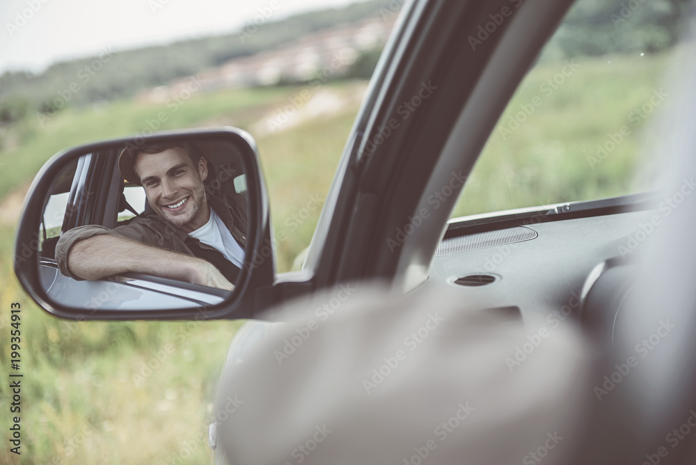 Cheerful guy is looking at side mirror of his car with smile. He is looking at camera with joy while sitting at seat of driver. Copy space in right side