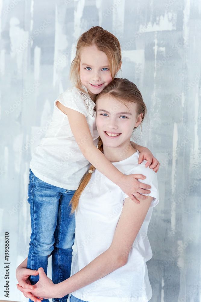 Two happy kids standing against grey textured wall background and embracing. Adorable pretty little girl hugging tight cute teenage girl, showing her love and care. Sisters having fun at home Two happy kids standing against grey textured wall background and embracing. Adorable pretty little girl hugging tight cute teenage girl, showing her love and care. Sisters having fun at home