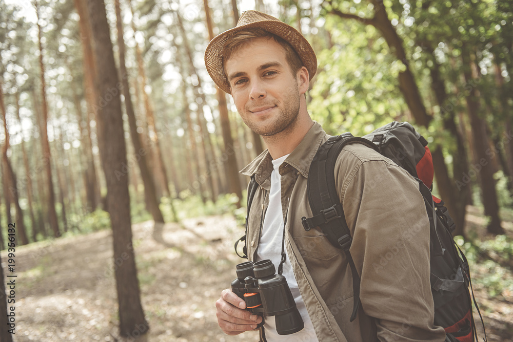 I feel good. Portrait of young positive tourist with backpack is standing in sunny forest and looking at camera with joy. He is holding binoculars
